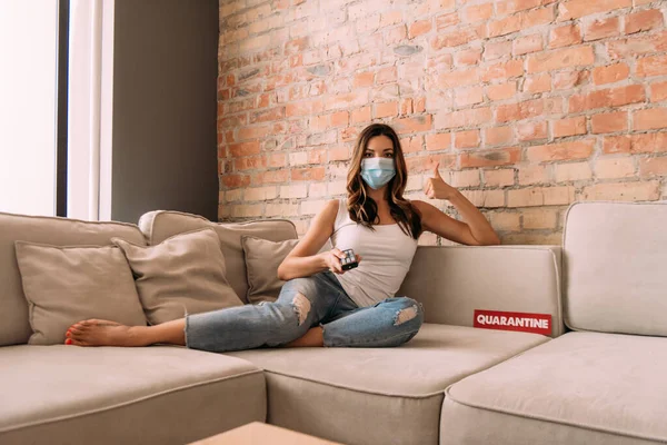 Woman in medical mask showing thumb up and holding remote controller while watching tv on sofa with quarantine sign — Stock Photo