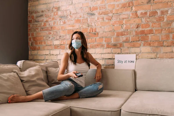 Beautiful girl in medical mask holding remote controller and watching tv on sofa with stay at home sign during self isolation — Stock Photo