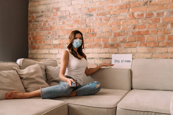 Frau in medizinischer Maske hält Fernbedienung in der Hand und schaut Fernsehen auf dem Sofa mit Bleibezeichen während der Selbstisolierung — Stockfoto