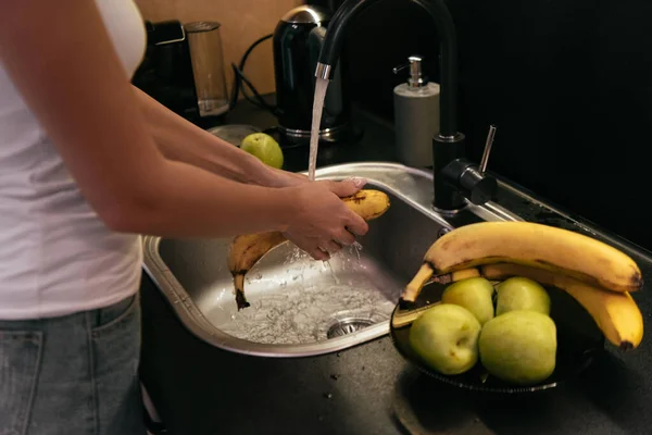 Обрезанный вид женщины, стирающей бананы и яблоки в раковине на кухне во время карантина — стоковое фото