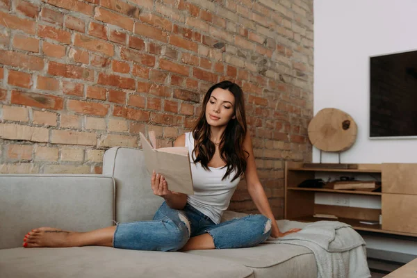 Положительная женщина читает книгу на диване во время самоизоляции — стоковое фото