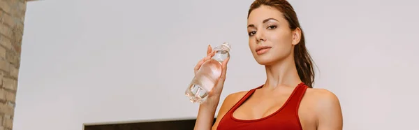 Attractive girl holding sports bottle and training at home during quarantine, panoramic crop — Stock Photo