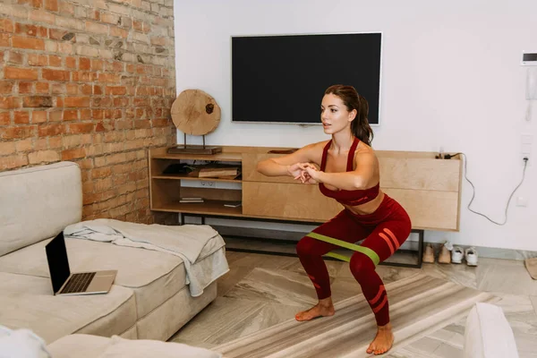 Schöne Sportlerin hockt mit Widerstandsband auf Fitnessmatte, trainiert online mit Laptop während der Selbstisolation — Stockfoto