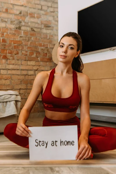 Beautiful sportswoman holding stay at home sign while sitting on fitness mat at home during self isolation — Stock Photo
