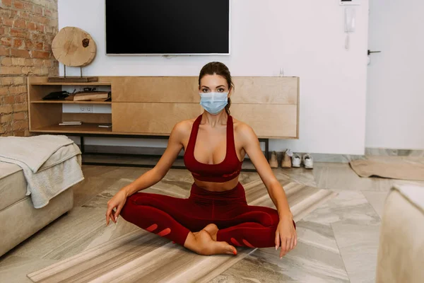 Chica en máscara médica sentado en pose de loto en esterilla de yoga en casa en cuarentena - foto de stock