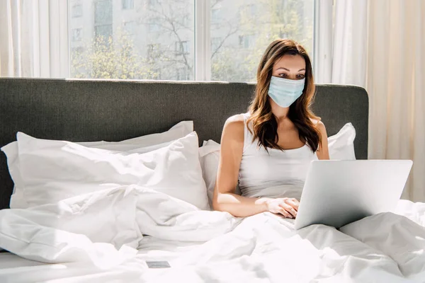 Freelancer femenina en máscara médica trabajando en portátil durante el autoaislamiento en la cama - foto de stock