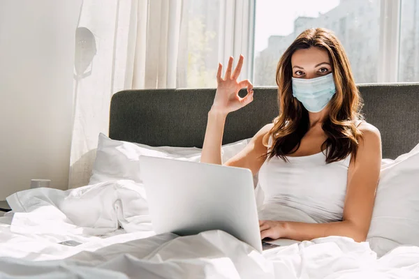 Freelancer femenina en máscara médica que muestra signo OK mientras trabaja en el ordenador portátil durante el autoaislamiento en la cama - foto de stock