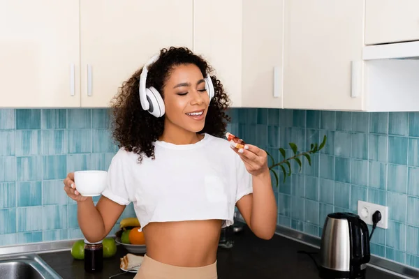 Felice ragazza afroamericana in cuffia wireless con pane tostato in mano con marmellata e tazza di caffè — Foto stock