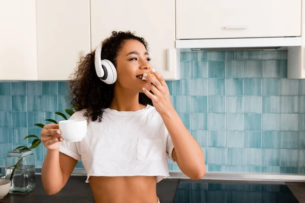 Happy african american girl in wireless headphones holding cup and eating toast bread with jam — Stock Photo