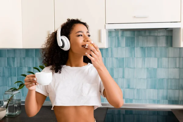 Heureux afro-américaine fille dans un casque sans fil tenant tasse et manger pain grillé avec confiture — Photo de stock