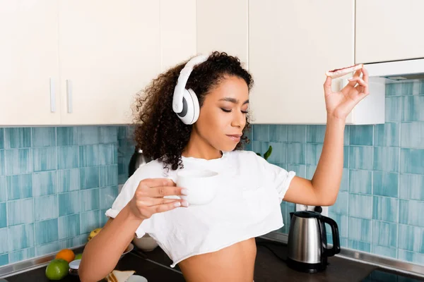 Atractiva chica afroamericana en auriculares inalámbricos sosteniendo pan tostado con mermelada y taza de café - foto de stock