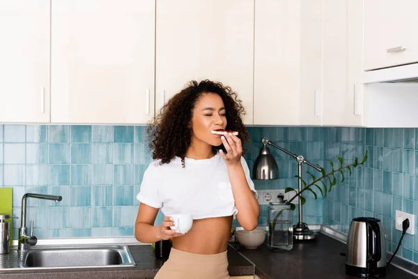 Fröhliches afrikanisch-amerikanisches Mädchen isst Toastbrot mit Marmelade und hält Tasse Kaffee in der Hand — Stockfoto