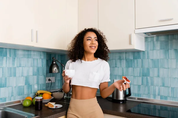 Menina americana africana alegre segurando pão torrado com geléia e xícara de café na cozinha — Fotografia de Stock