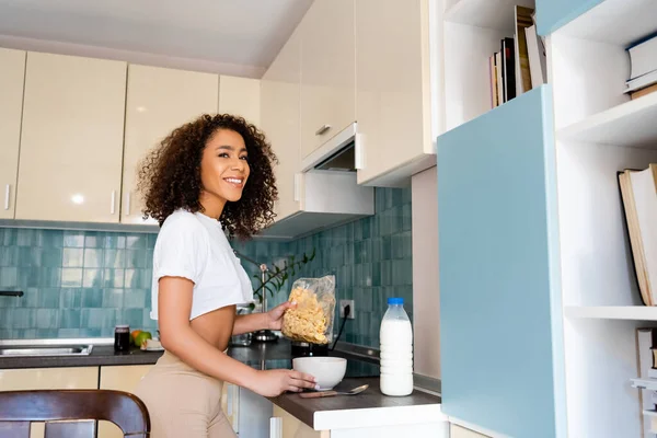 Mujer afroamericana feliz sosteniendo paquete con copos de maíz cerca de tazón y botella con leche - foto de stock