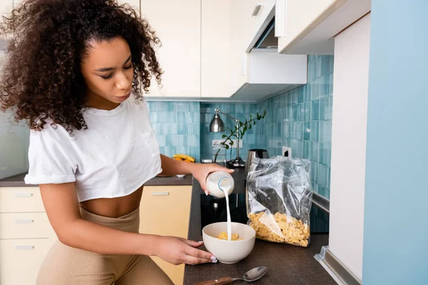 Mujer afroamericana vertiendo leche en un tazón con copos de maíz - foto de stock