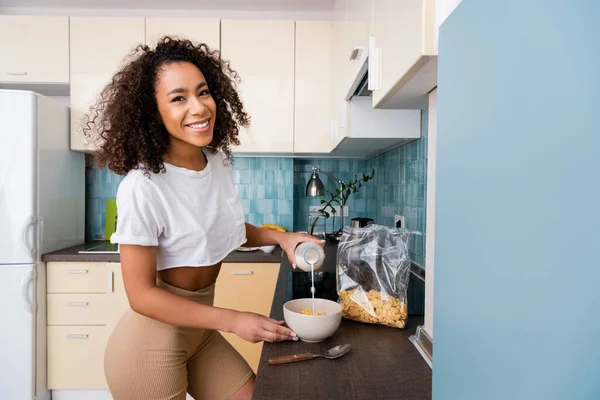 Mujer afroamericana feliz vertiendo leche en un tazón con copos de maíz - foto de stock