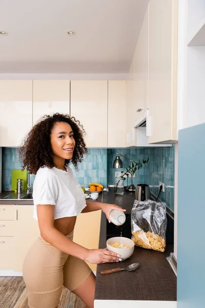 Cheerful african american woman pouring milk in bowl with cornflakes — Stock Photo