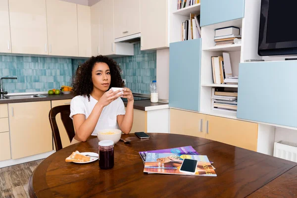 Attraente donna afro-americana in possesso di tazza con caffè vicino gustosa colazione e smartphone con schermo bianco — Foto stock