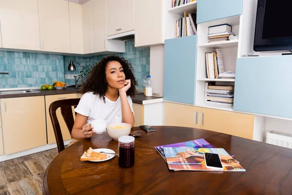 Encaracolado Africano americano mulher segurando xícara com café perto de café da manhã saboroso e smartphones com tela em branco — Fotografia de Stock