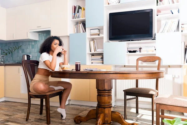 Frisé afro-américaine femme boire du café près de savoureux petit déjeuner et tv avec écran blanc — Photo de stock