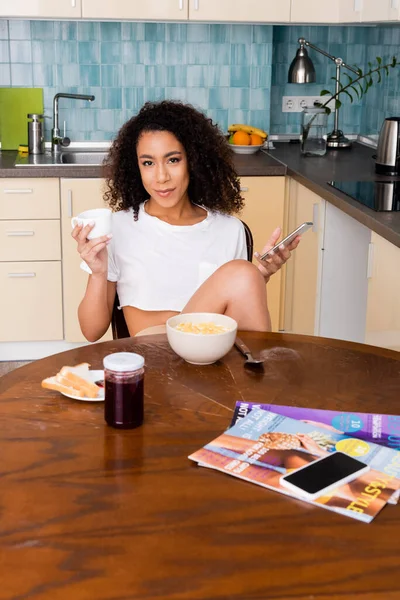 Attractive african american woman holding smartphone and cup near tasty breakfast and gadget with blank screen — Stock Photo