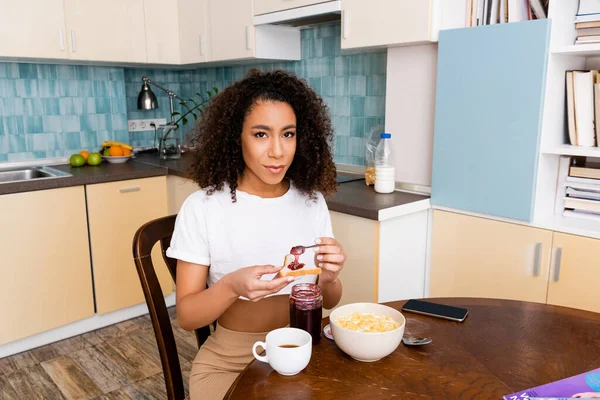 Donna riccia africana americana che tiene cucchiaio con marmellata dolce vicino al pane tostato e smartphone con schermo bianco — Foto stock