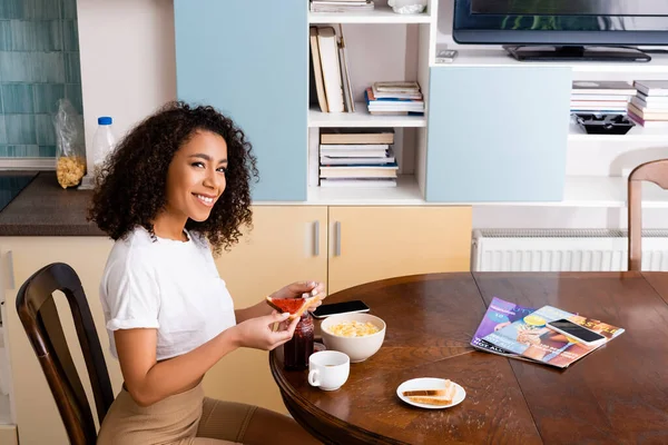 Happy and curly african american woman holding spoon near toast bread and corn flakes in bowl — Stock Photo