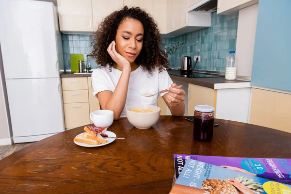 Femme afro-américaine tenant cuillère avec des flocons de maïs près du bol, pain grillé, confiture et tasse avec café sur la table — Photo de stock