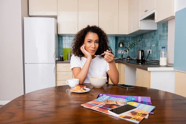 Femme afro-américaine tenant cuillère avec flocons de maïs près de tasse avec café et smartphone avec écran blanc sur la table — Photo de stock