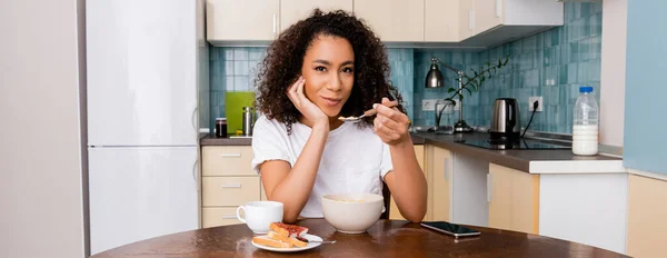 Récolte panoramique de femme afro-américaine tenant cuillère avec des flocons de maïs près de tasse avec café et smartphone avec écran blanc sur la table — Photo de stock