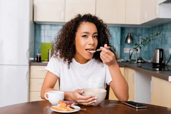 Mujer afroamericana comiendo hojuelas de maíz cerca de la taza con café, pan tostado y teléfono inteligente con pantalla en blanco en la mesa - foto de stock