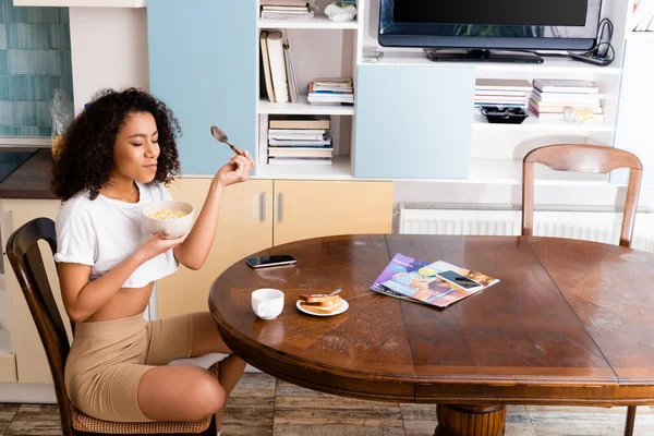 Attrayant afro-américaine femme tenant bol avec savoureux flocons de maïs et cuillère près de gadgets avec écran blanc — Photo de stock