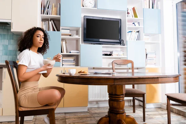 Femme afro-américaine tenant bol avec de savoureux flocons de maïs et cuillère dans la cuisine moderne — Photo de stock