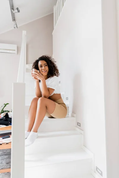 Fröhliche afrikanisch-amerikanische Frau sitzt mit einer Tasse Kaffee auf der Treppe — Stockfoto
