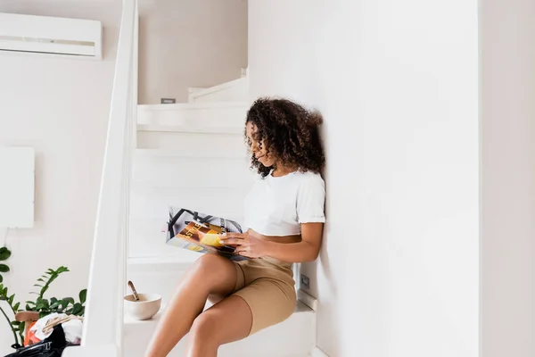 Mujer afroamericana sentada en escaleras con revista cerca de tazón con hojuelas de maíz - foto de stock