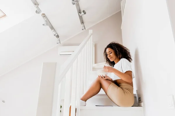 Mujer afroamericana feliz sentada en las escaleras y sosteniendo tazón y cuchara - foto de stock