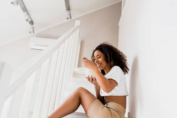 Happy african american woman sitting on stairs and holding bowl with breakfast — Stock Photo