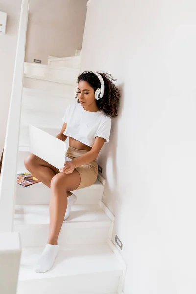 Afro-américain freelance dans les écouteurs sans fil assis sur les escaliers et en utilisant un ordinateur portable à la maison — Photo de stock