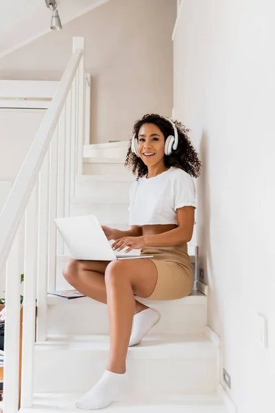 Feliz afroamericano freelancer en auriculares inalámbricos sentado en las escaleras y utilizando el ordenador portátil - foto de stock