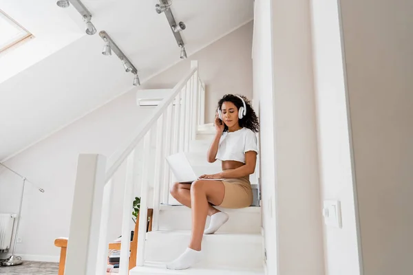 Bastante afroamericano freelancer en auriculares inalámbricos sentado en las escaleras y utilizando el ordenador portátil - foto de stock