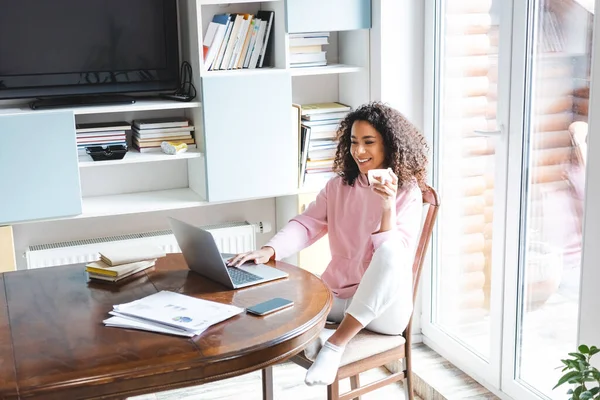 Heureux afro-américain pigiste tenant tasse tout en utilisant un ordinateur portable à la maison — Photo de stock