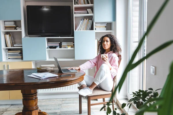 Selektiver Fokus afrikanisch-amerikanischer Freelancer, der Tasse hält, während er Laptop zu Hause benutzt — Stockfoto