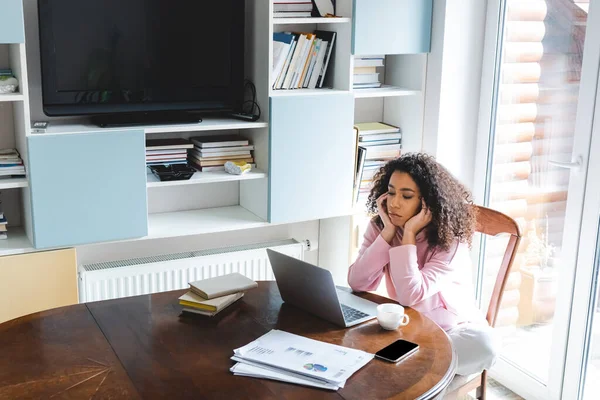 Upset african american freelancer looking at laptop near smartphone with blank screen, books and cup — Stock Photo