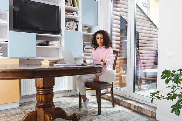 Atraente freelancer afro-americano olhando para documentos perto de laptop, livros e xícara — Fotografia de Stock