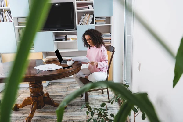 Selective focus of attractive african american freelancer looking at documents near gadgets, books and cup — Stock Photo