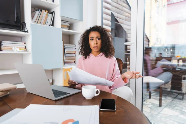 Selective focus of attractive african american freelancer holding documents and gesturing near gadgets and cup — Stock Photo
