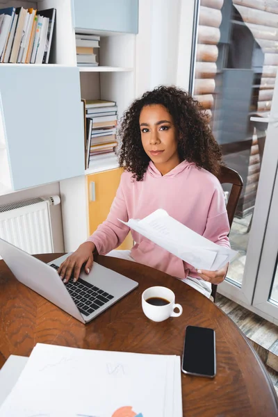 Rizado afroamericano freelancer celebración de documentos cerca de la computadora portátil, teléfono inteligente con pantalla en blanco y taza — Stock Photo