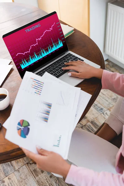 Cropped view of african american freelancer holding charts and graphs near laptop with  trading on screen — Stock Photo