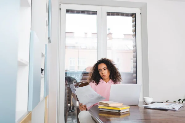 Selective focus of curly african american freelancer looking at documents near laptop, books and cup — Stock Photo