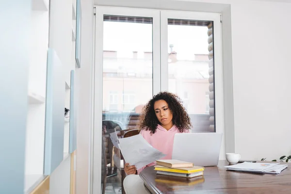 Foyer sélectif de freelance afro-américaine frisée regardant des documents près d'un ordinateur portable, livres et tasse — Photo de stock